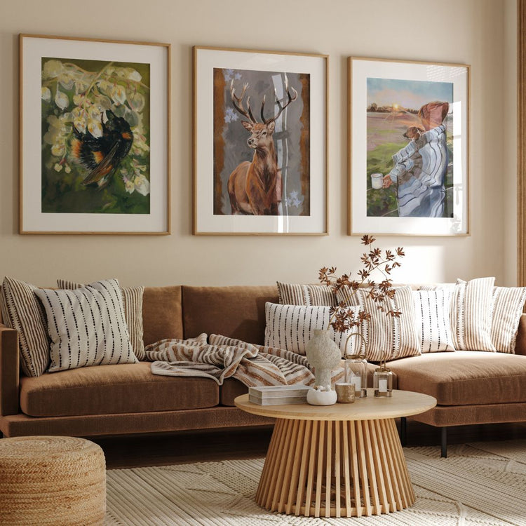 Gallery wall with framed fine art prints of a bee on blossoms, a stag, and a countryside sunrise with a woman and her dog. Cozy living room with modern rustic decor, warm tones, and nature-inspired artwork. Perfect for home interiors and nature lovers.