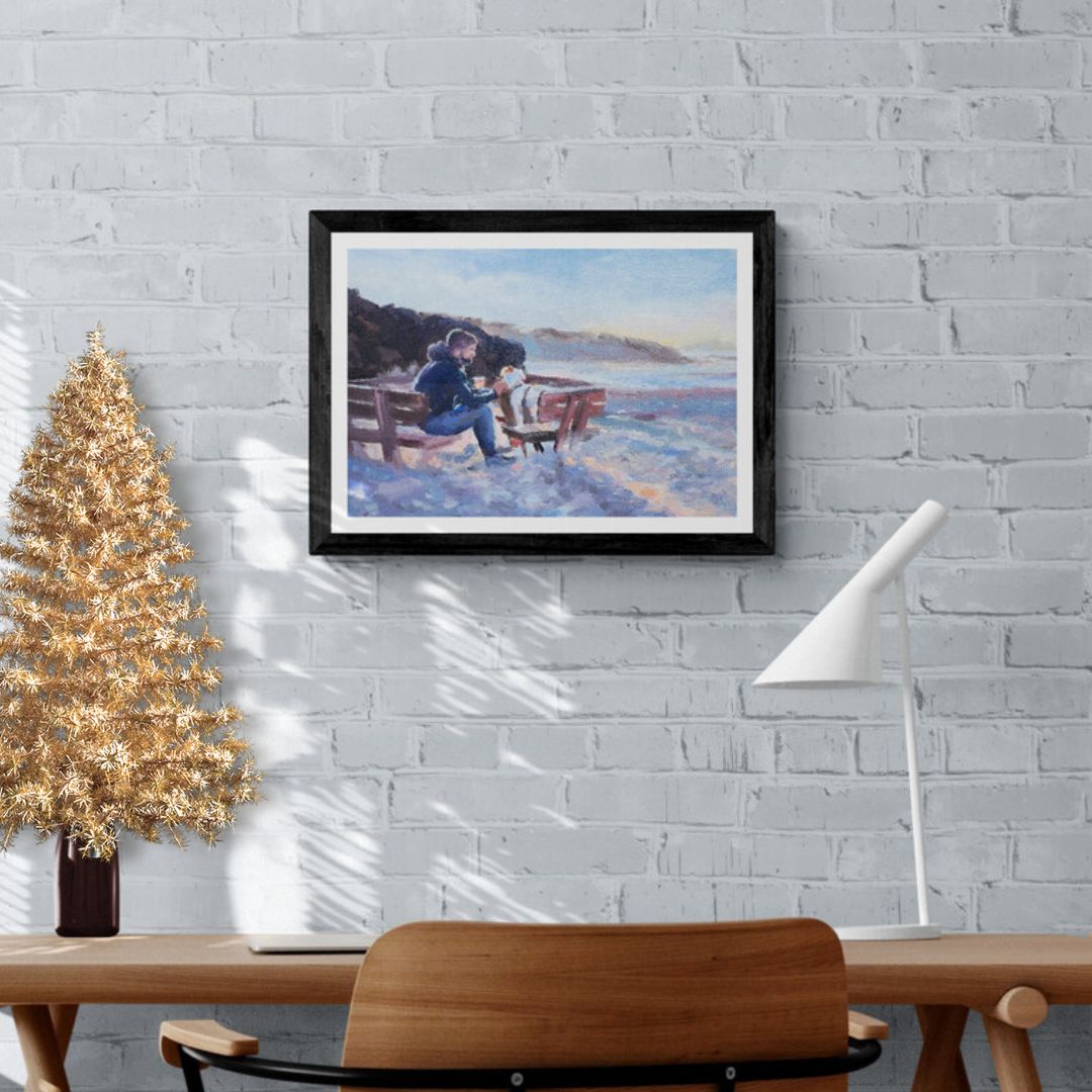 A framed artwork depicting a scene of a man and a dog sitting on a seaside bench, with the dog being fussed by the man. The artwork is displayed on a wall above a wooden table and a gold-colored Christmas tree.