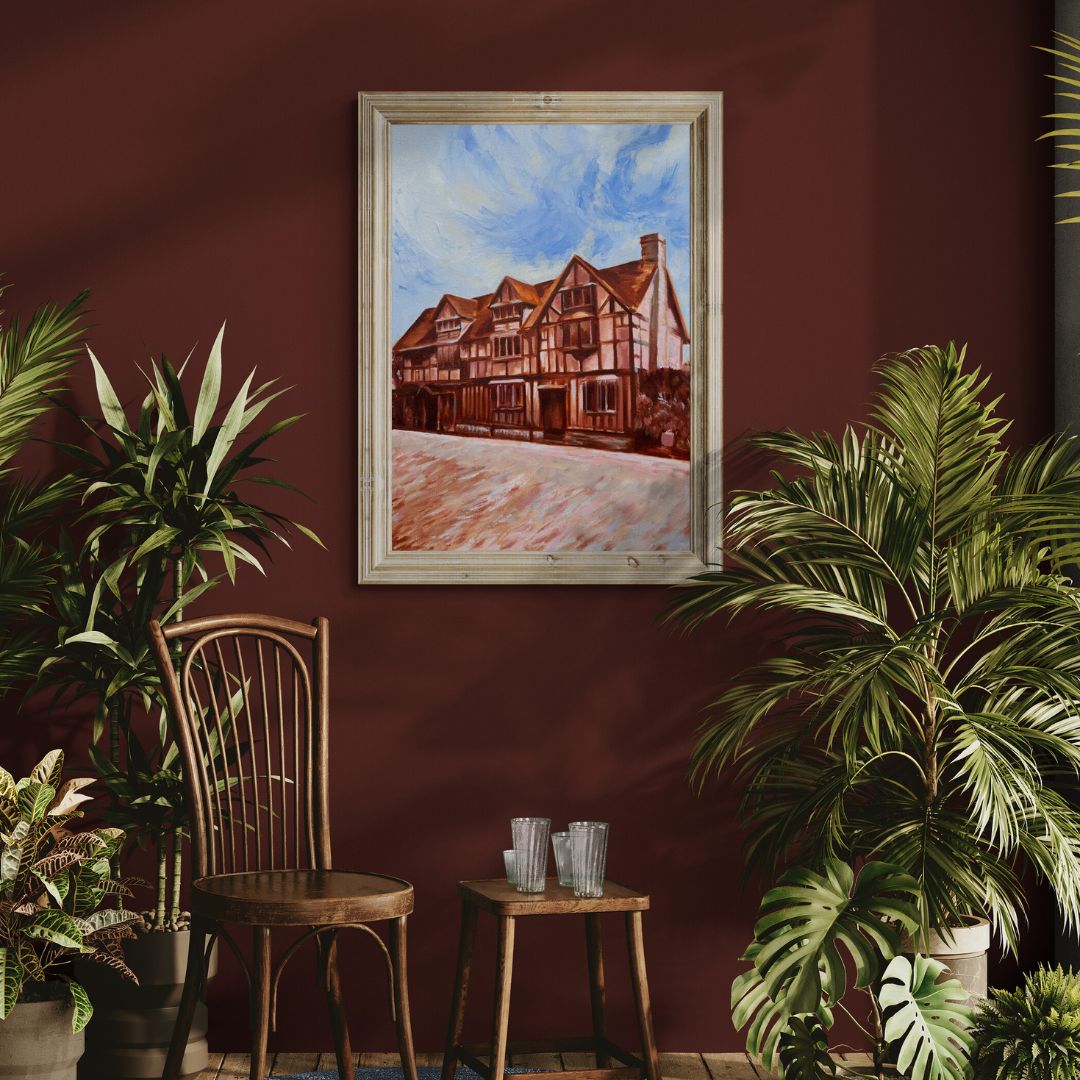 Original oil painting of Shakespeare’s Birthplace in Stratford-upon-Avon by Lorraine Field, framed in natural wood frame mock-up and displayed above a rustic chair with plants against a deep red wall.