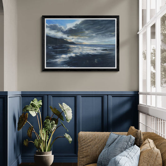 Framed art print of coastal scene on a wall with a large glazed window, plant and sofa below.