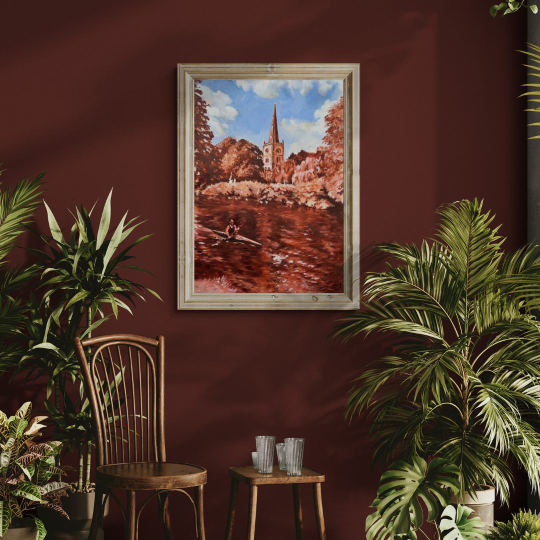 Original oil painting of Holy Trinity Church in Stratford-upon-Avon by Lorraine Field, framed in natural wood and displayed above a rustic chair with plants against a deep red wall.