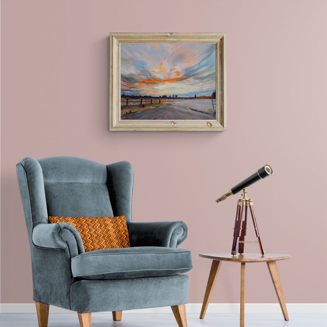 Elegant reading nook with a framed fine art painting of a Warwickshire sunrise. Features a blue velvet armchair, wooden side table with a vintage telescope, and a soft pink wall. Perfect cozy interior with nature-inspired artwork and warm tones.