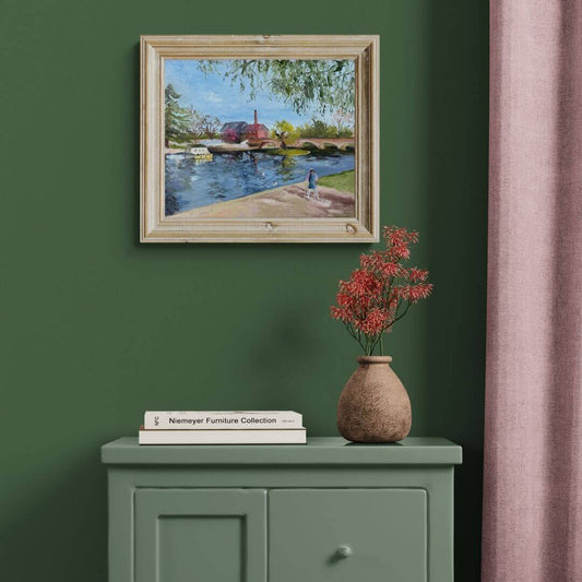 Original oil painting of a young girl walking beside the River Avon at Bridgefoot in Stratford-upon-Avon, displayed in a light wood frame above a sage green cabinet. The artwork captures the peaceful riverside with a stone bridge, willow trees and boats beneath a blue sky.