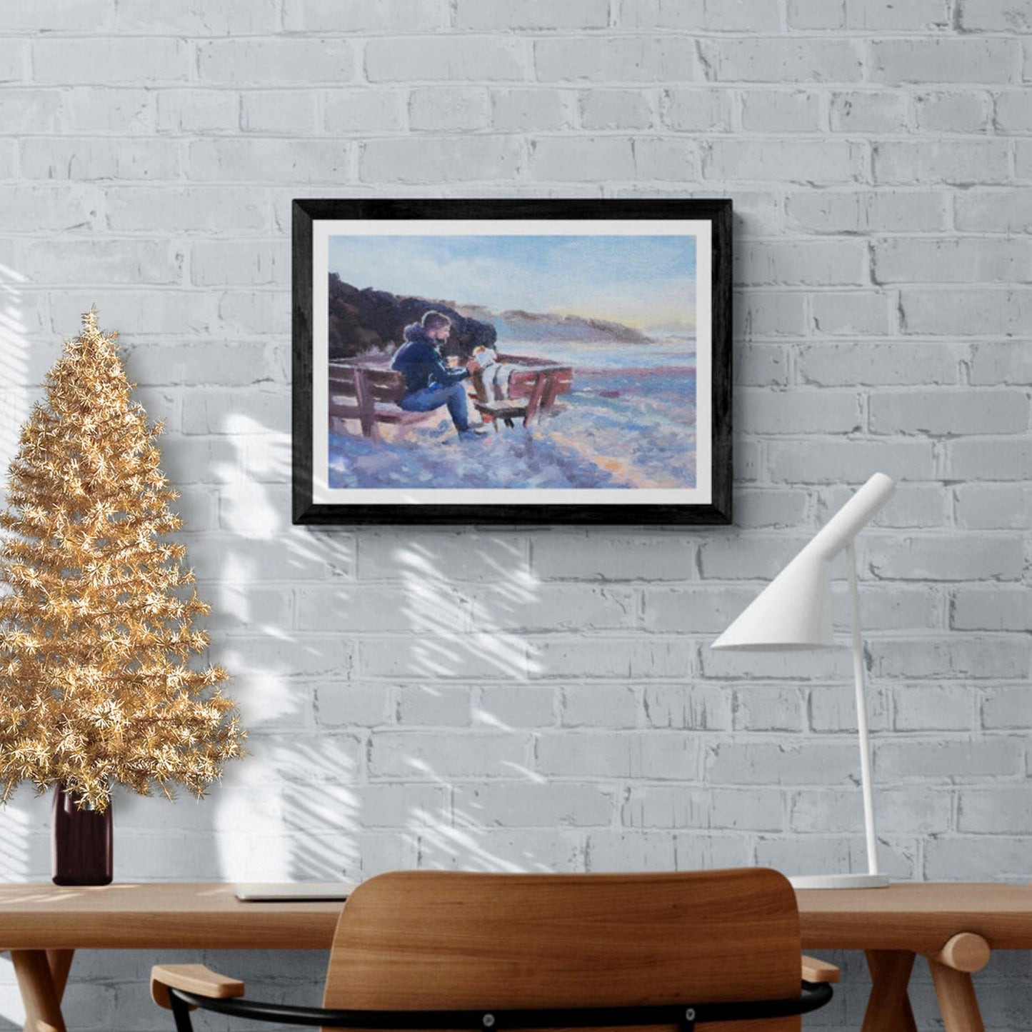 A framed artwork depicting a scene of a man and a dog sitting on a seaside bench, with the dog being fussed by the man. The artwork is displayed on a wall above a wooden table and a gold-coloured Christmas tree.