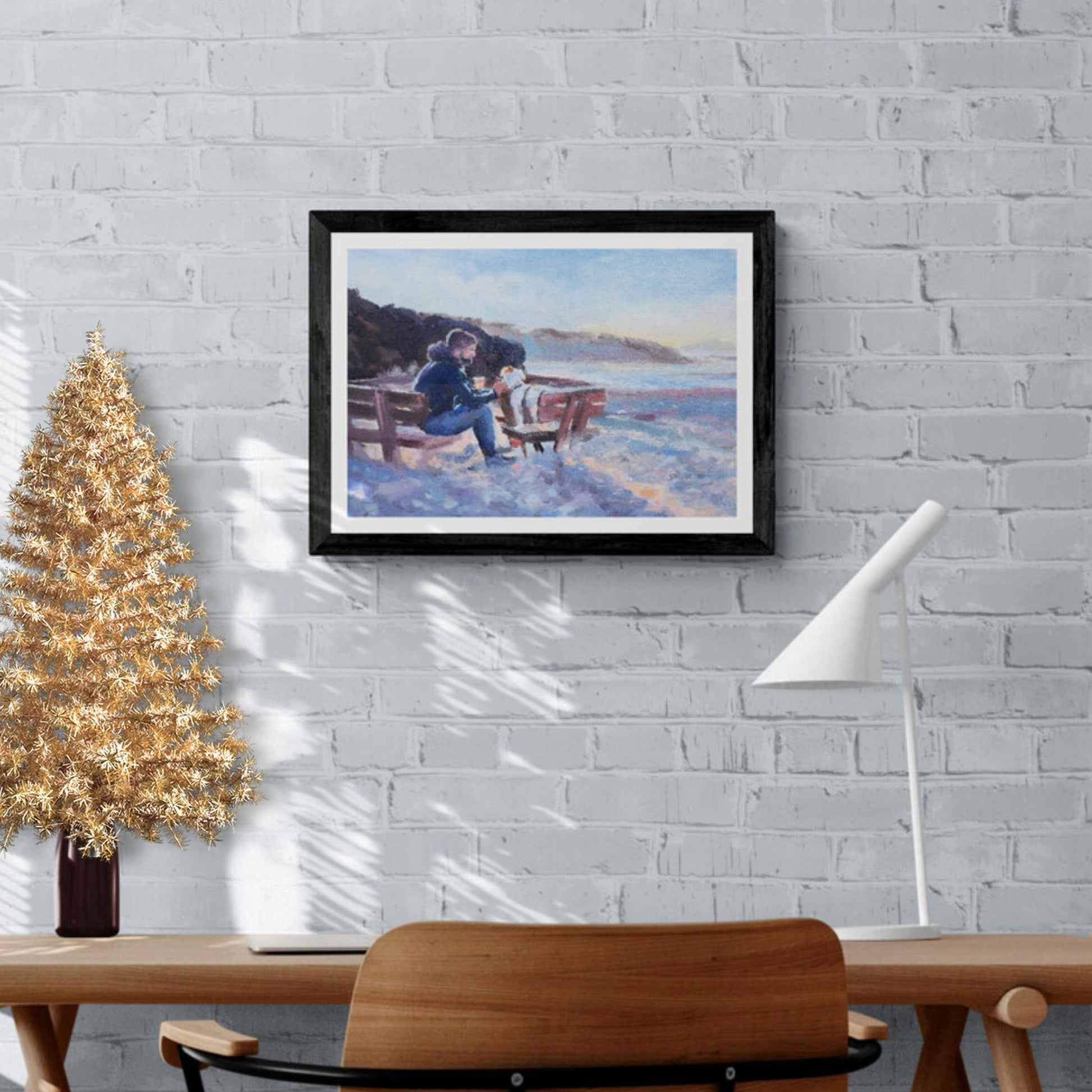 A framed artwork depicting a scene of a man and a dog sitting on a seaside bench, with the dog being fussed by the man. The artwork is displayed on a wall above a wooden table and a gold-coloured Christmas tree.