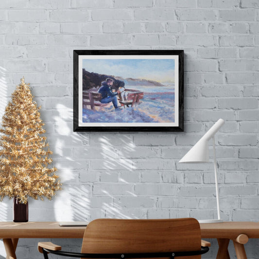 A framed artwork depicting a scene of a man and a dog sitting on a seaside bench, with the dog being fussed by the man. The artwork is displayed on a wall above a wooden table and a gold-coloured Christmas tree.