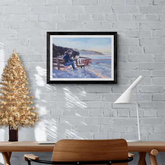 A framed artwork depicting a scene of a man and a dog sitting on a seaside bench, with the dog being fussed by the man. The artwork is displayed on a wall above a wooden table and a gold-colored Christmas tree.