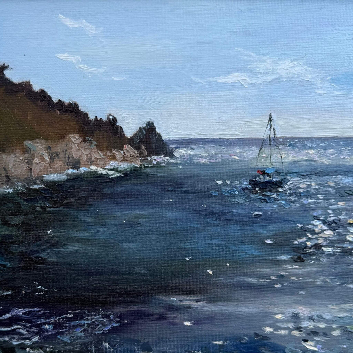 Close up view of Lamorna Cove with sailing boat oil painting by Lorraine Field, on sparkling blue water in a pale wooden frame.