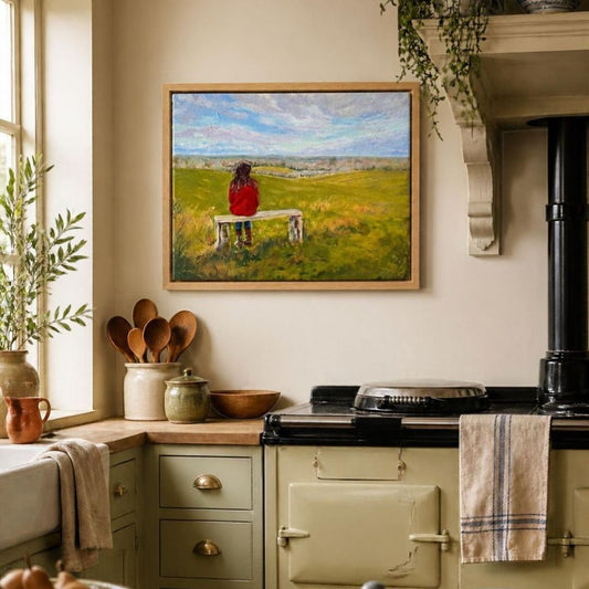 If Fields Could Talk’ original oil painting by Lorraine Field in a natural wood frame in a country kitchen, depicting a woman in red seated on a bench overlooking green fields.
