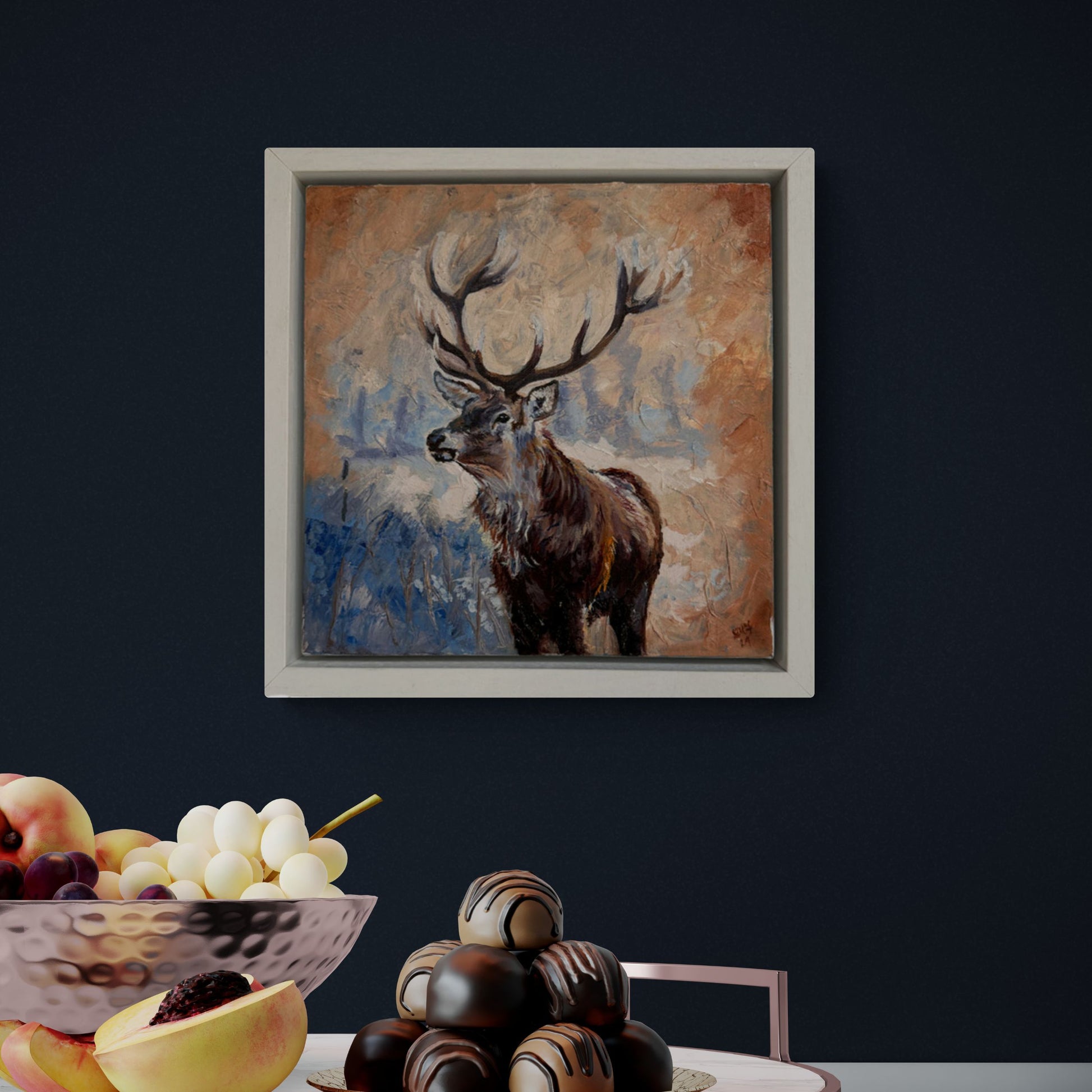 Framed original stag oil painting on a deep blue wall above a table with fruit and chocolates.