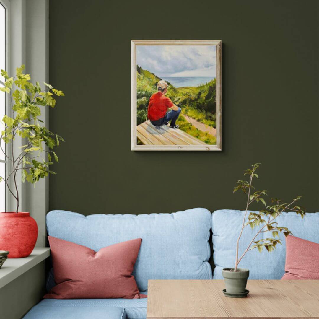 Just Taking It In oil painting by Lorraine Field displayed above a pale blue sofa on a dark olive-green wall, with pink cushions and indoor plants.

