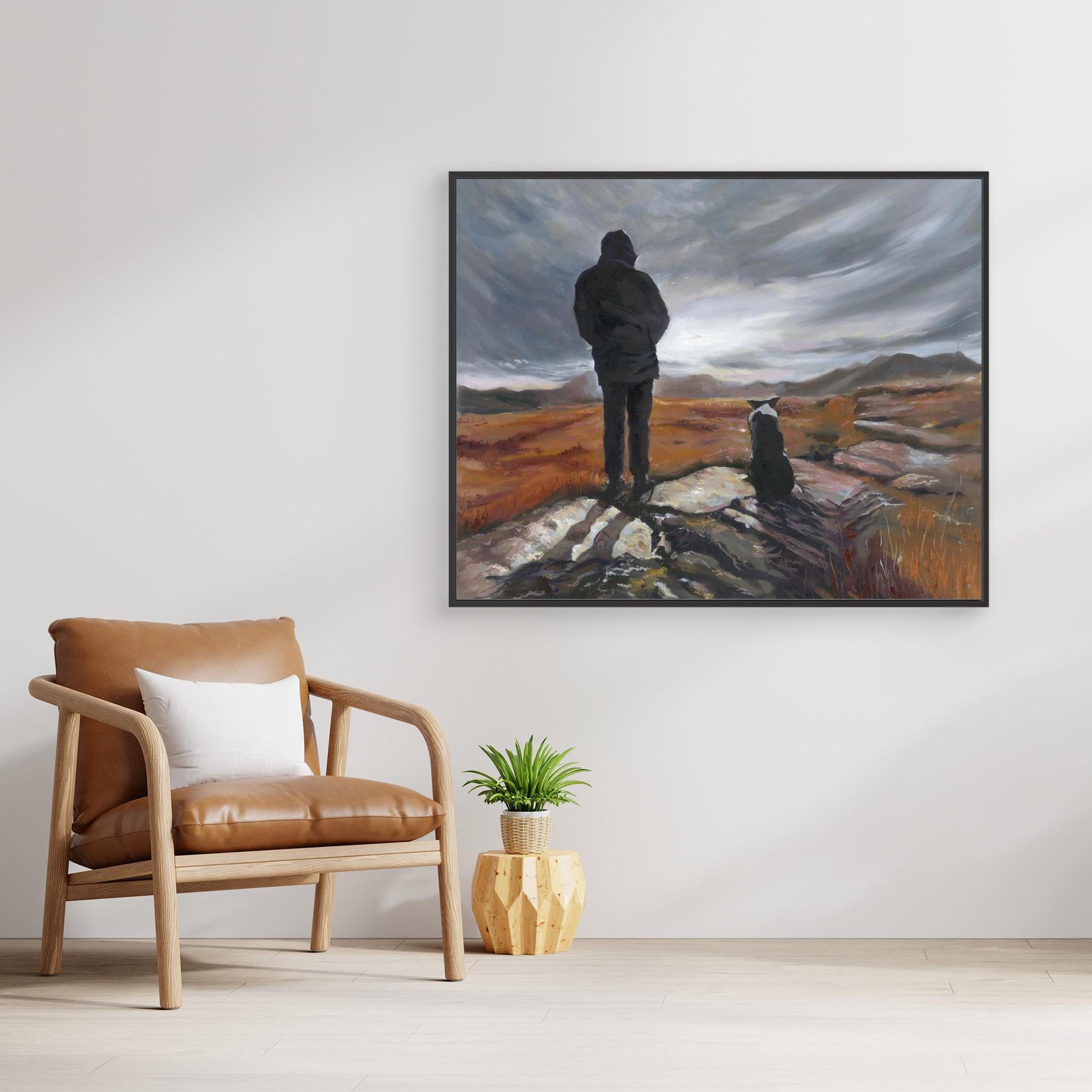 Framed canvas art print of a man and Border Collie dog under grey sky landscape, above a brown leather armchair with a white pillow.
