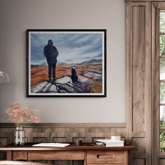 Framed print of ‘Man and His Border Collie’ artwork in a sleek black frame, styled above a rustic wooden desk with soft, homely decor.