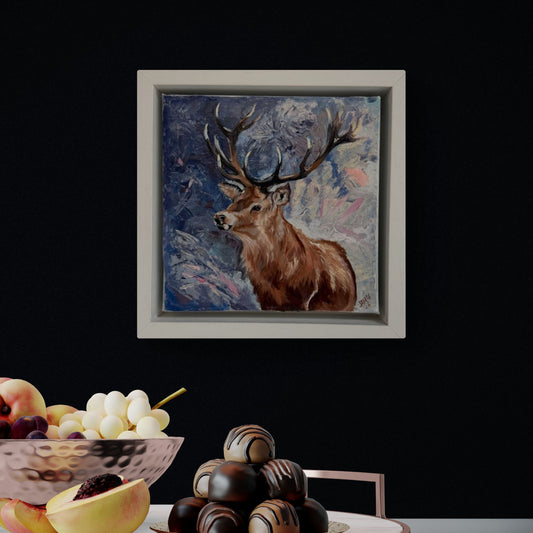 Framed deer oil painting on a dark blue wall with a fruit bowl and chocolate tower in the foreground.