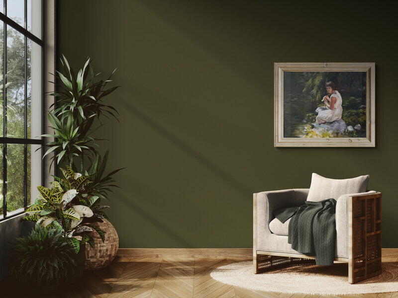 A styled interior scene with a dark olive green wall, a framed oil painting "Stillness Among the Leaves" by British Artist Lorraine Field hanging on the wall, a light upholstered armchair with a dark green throw blanket, a round rug, and potted plants near a window.