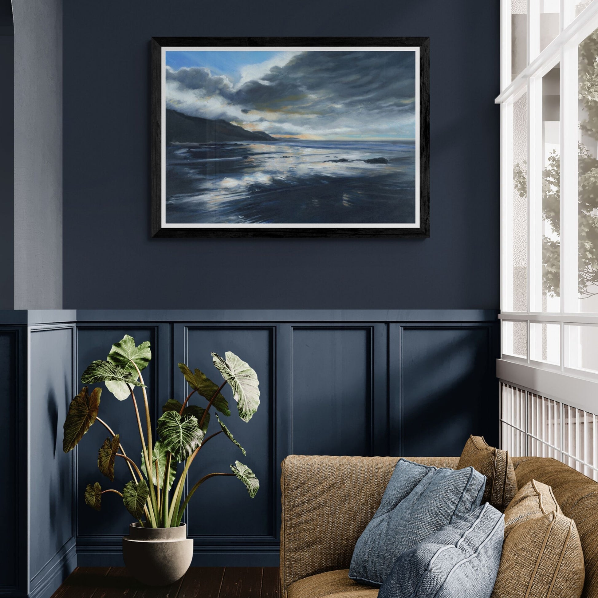 Framed art print of coastal scene on a wall with a large glazed window, plant and sofa below.