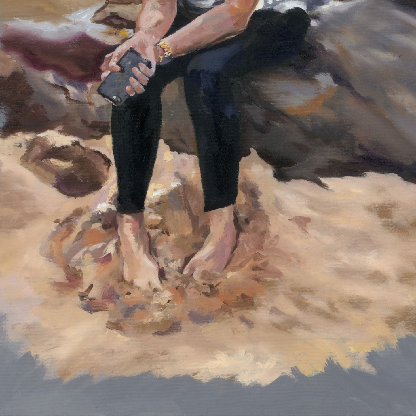 Young lady sitting on a rock with her feet buried in sand, wearing black trousers and holding a mobile phone.