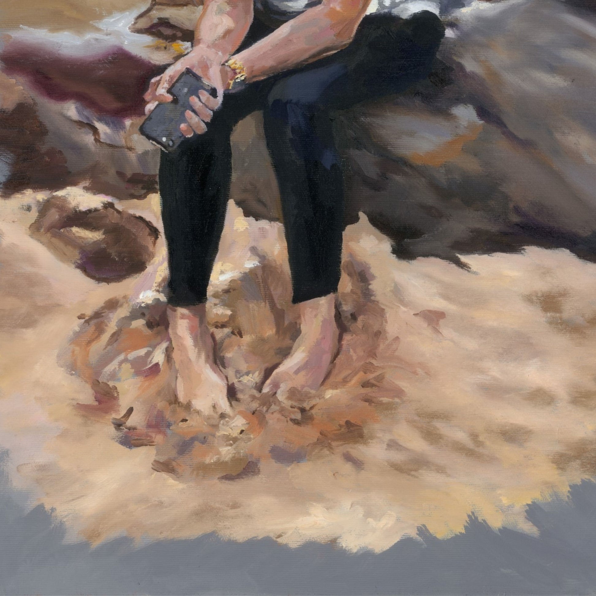 Young lady sitting on a rock with her feet buried in sand, wearing black trousers and holding a mobile phone.