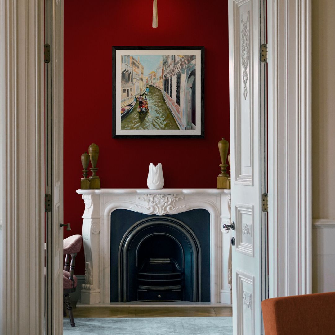 Decorative interior with a white fireplace and framed Venice artwork on a deep red wall.
