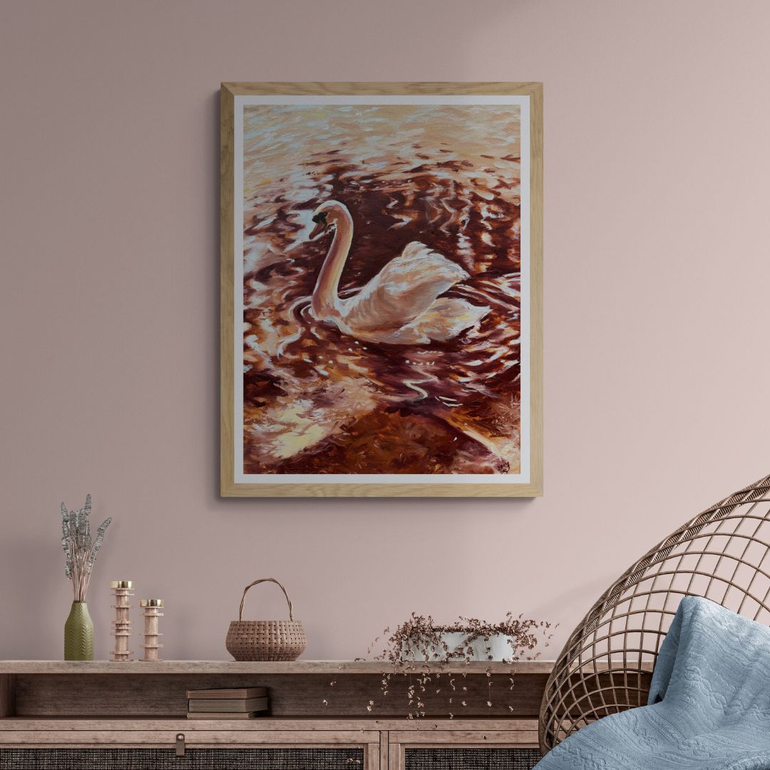“Becoming Grace” swan fine art print in a natural wood frame, styled on a soft blush pink wall above a wooden console table. Warm copper reflections and peaceful movement create a calming atmosphere.
