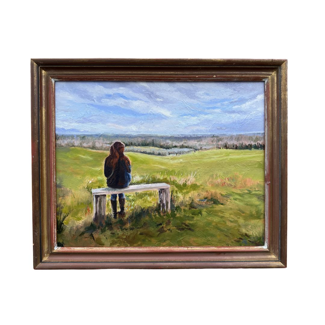 Original oil painting in mock up frame, titled If These Fields Could Talk by Lorraine Field, showing a lone woman sitting on a bench overlooking rolling countryside.