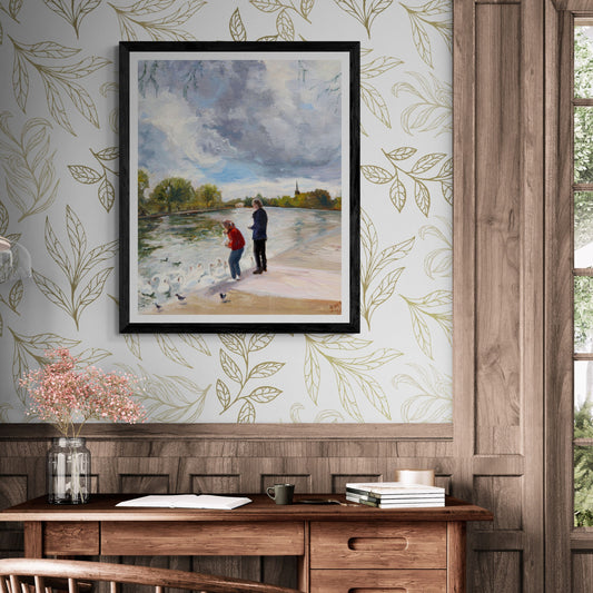 Framed Swans in Stratford fine art print by Lorraine Field Art displayed above a wooden desk, featuring people feeding swans beside the River Avon in Stratford-upon-Avon.
